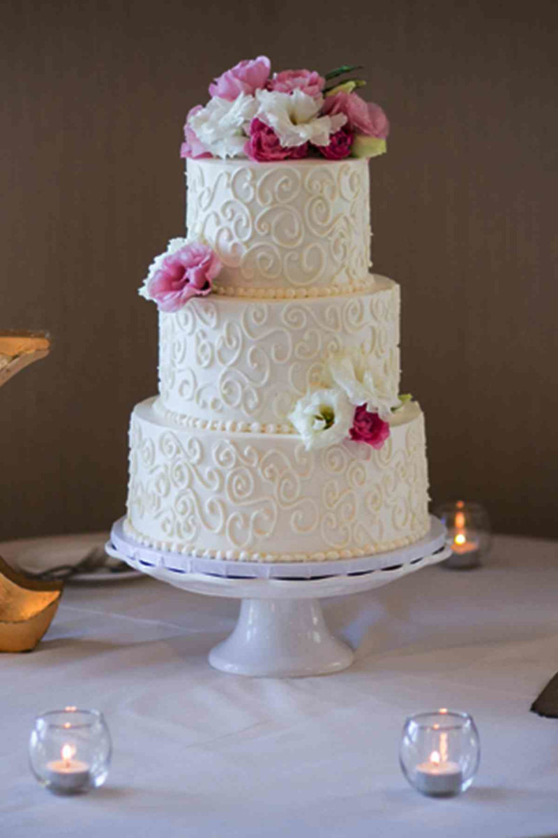 Simple three-layered white wedding cake with swirls, pearls and pink flowers.